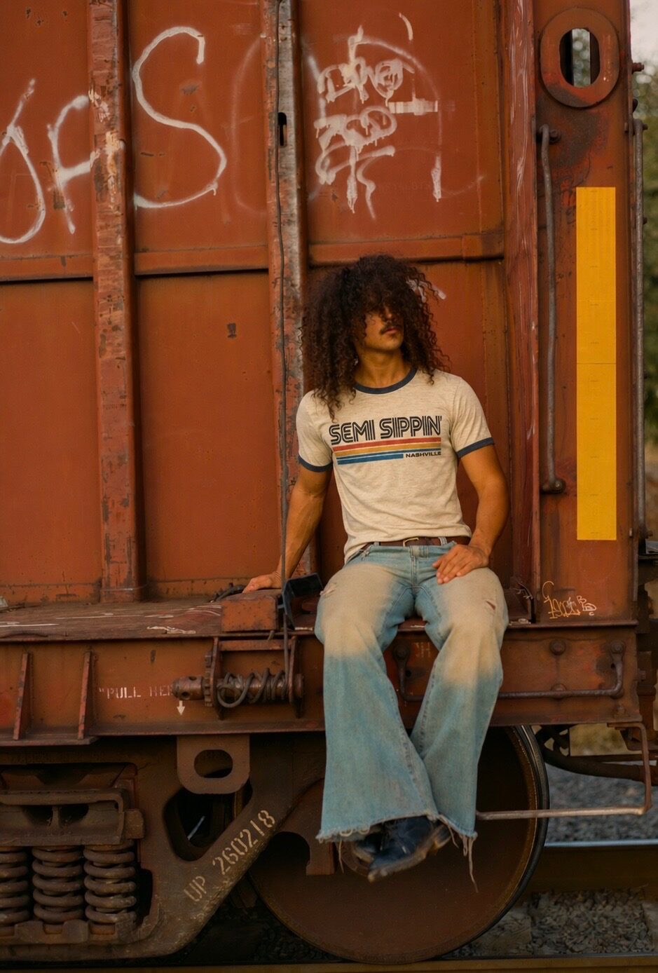 A man in a ringer tee sitting on the coupling of a rust-red freight car